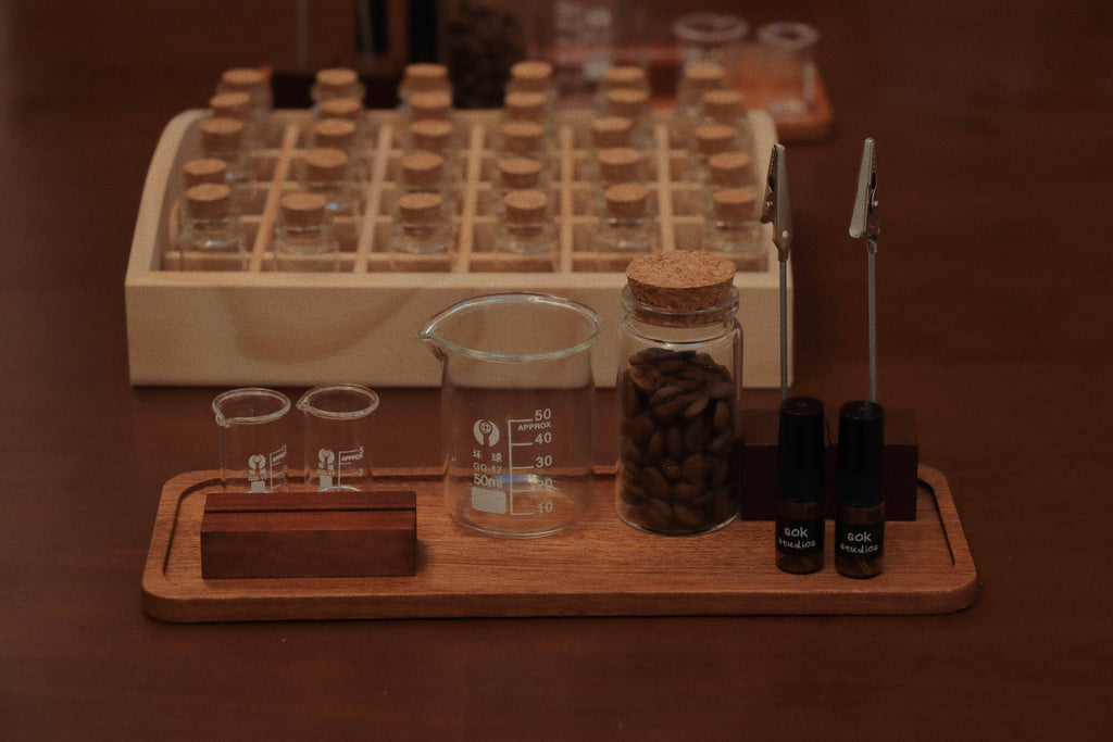 Perfume blending tools including beakers, coffee beans in a jar, scent clips, and SOK Studios sample bottles on a wooden tray, with empty vials in the background